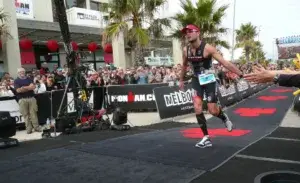 A powerful scene at the Ironman triathlon finish line, capturing the moment an athlete crosses after completing the swim, bike, and run. The atmosphere includes cheering fans, Ironman branding, and a race clock under the finish arch