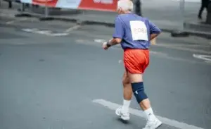 Senior man running after knee replacement during a road race, wearing a knee brace for support.