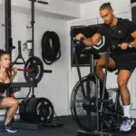 Man using an exercise bike with arm workout in a home gym next to a woman lifting weights.