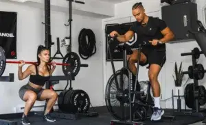 Man using an exercise bike with arm workout in a home gym next to a woman lifting weights.